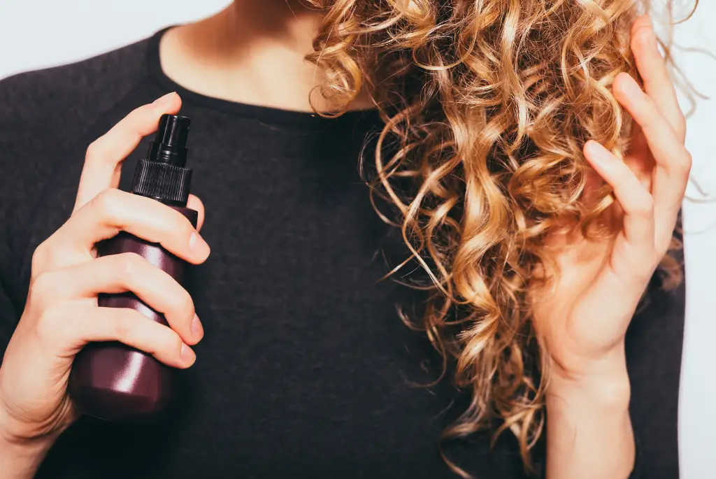 Person applying hair spray to curly blonde hair for styling and volume